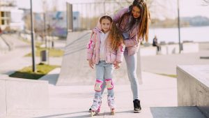 Full body cheerful young woman supporting daughter wearing protection pads while roller skating on concrete ramp during active leisure in sunny spring day